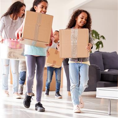 Mother and three children carrying big brown cardboard boxes out of the house smiling. 