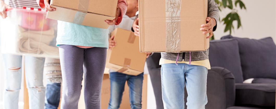 Mother and three children carrying big brown cardboard boxes out of the house smiling. 